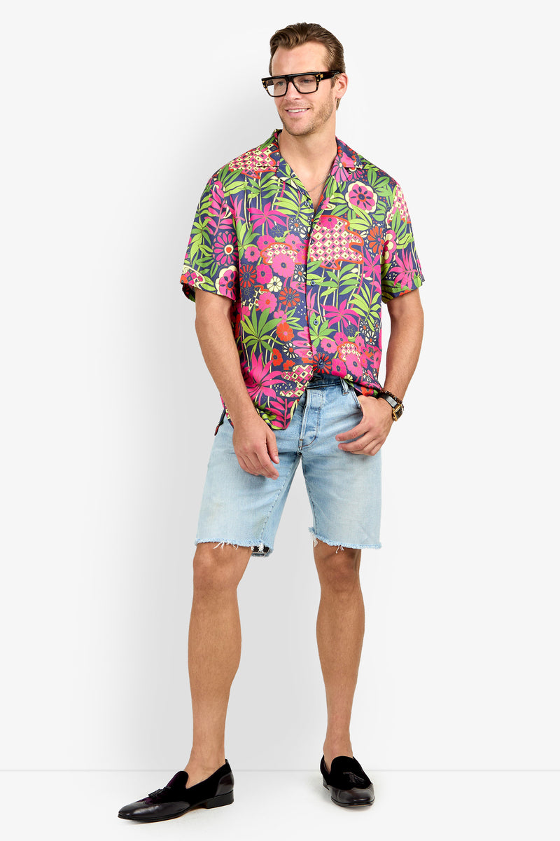 A man wearing the Felix Camp Shirt, light denim shorts, black loafers, and glasses stands smiling with one hand in his pocket against a plain white background.