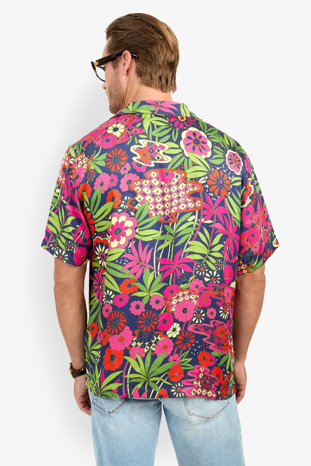A man wearing glasses and the Felix Camp Shirt, a colorful short-sleeve floral shirt with green, pink, red, and yellow patterns, stands facing away. He also wears light blue jeans.