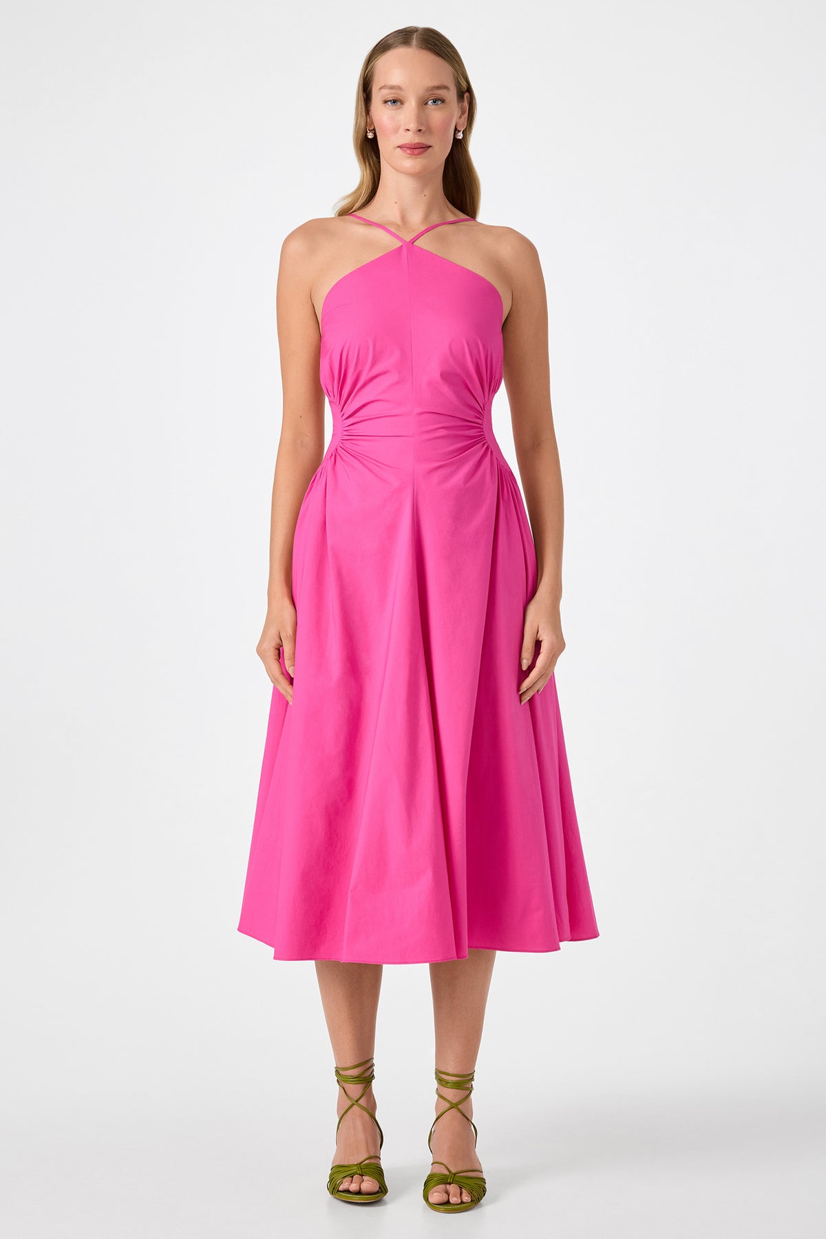 A woman with long hair faces forward, wearing the bright pink Kata Shirred Waist Midi Dress with a halter neck, sleeveless design, and flared skirt. She pairs it with green strappy sandals against a plain white background.