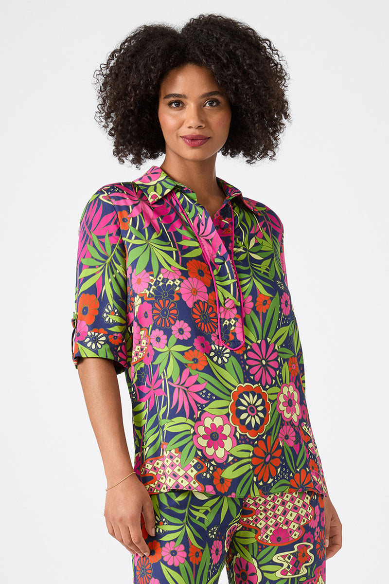 A woman with curly hair and a slight smile wears the Portrait 2 Top in Fumi Floral, featuring bold pink, red, green, and yellow flowers with leafy designs on a dark background.