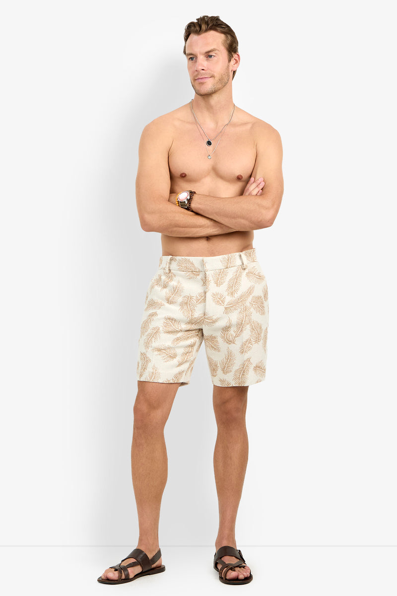 A shirtless man wearing the Lawrence Short, patterned in beige, with dark sandals and necklaces stands with arms crossed against a plain white background.