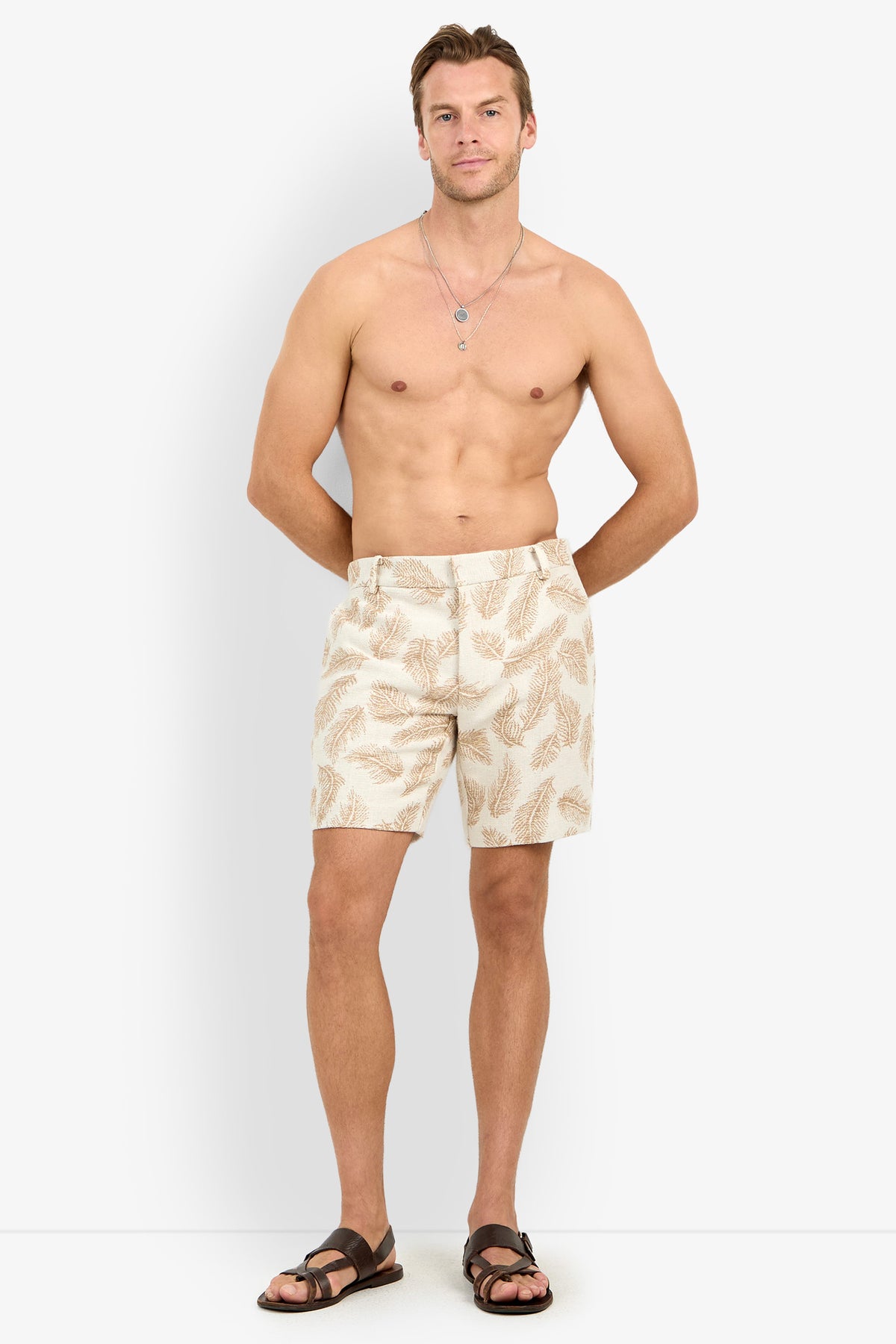 A barefoot man with short hair wears the Lawrence Short in a beige leaf pattern and brown sandals, standing shirtless with his hands in his pockets against a plain white background.