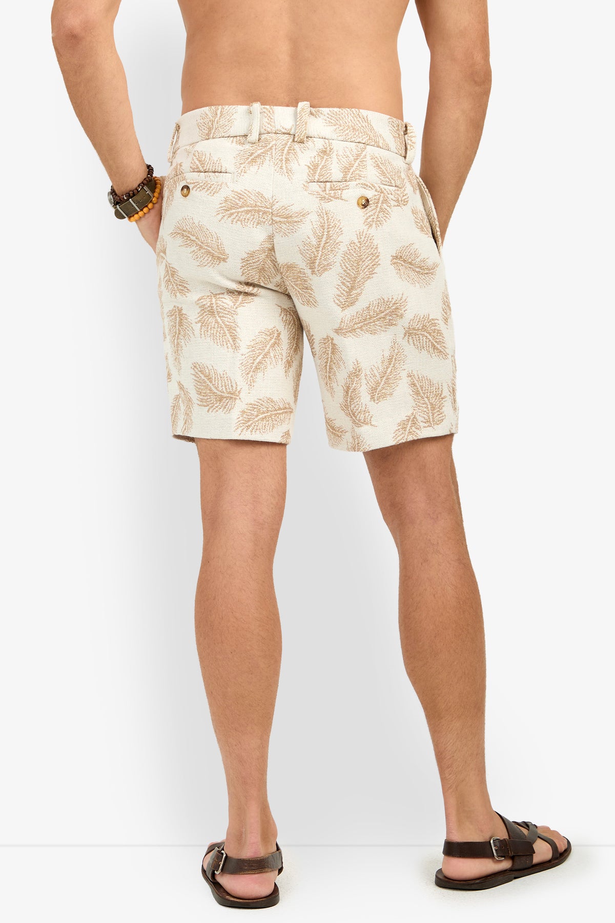 A man stands facing away against a white background, wearing Lawrence Short in a tan palm-leaf print, paired with brown sandals and a beaded bracelet, shirtless.