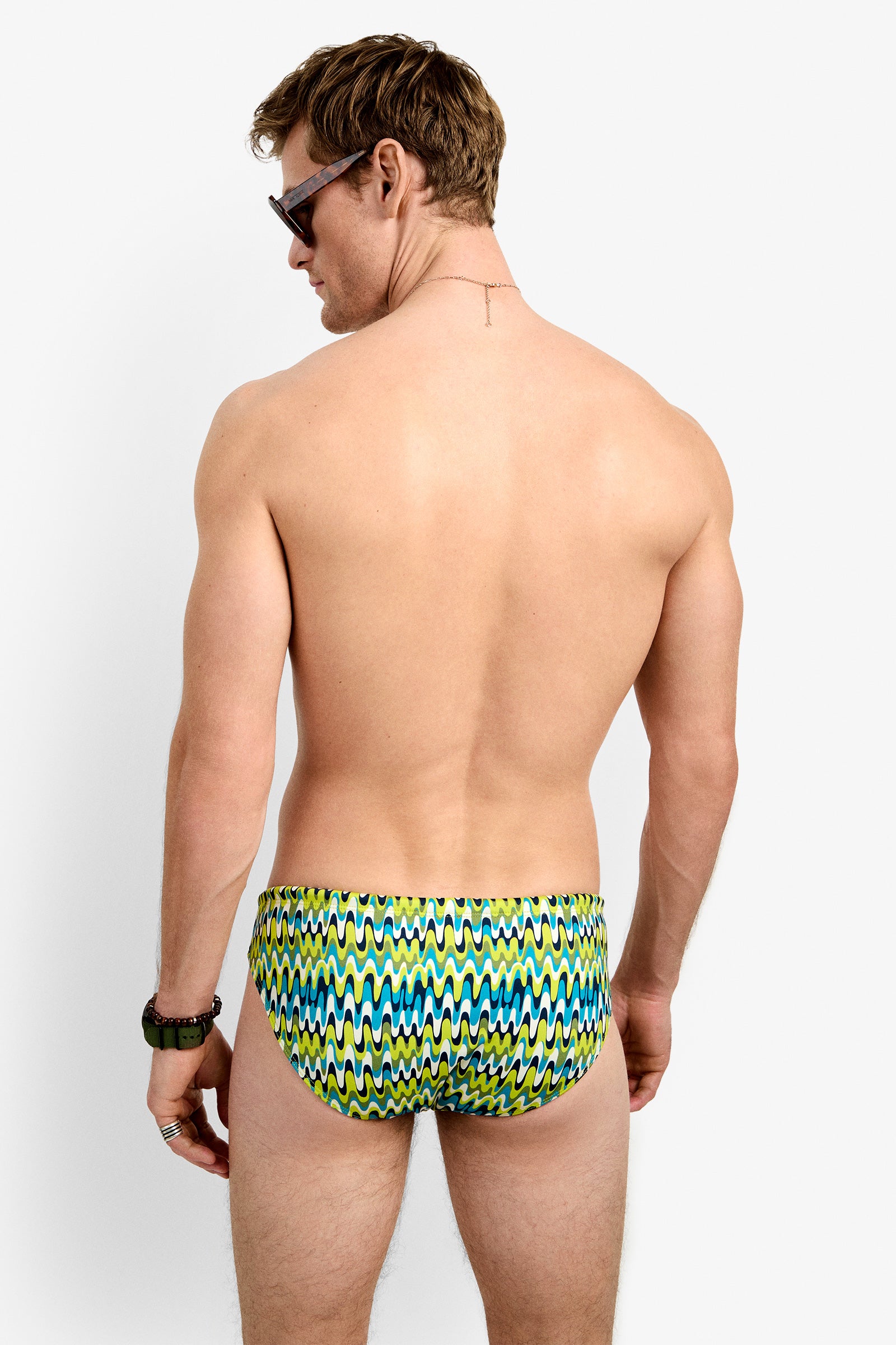 A man with short hair, sunglasses, a gold necklace, and a black wristwatch is shown from the back against a white background, wearing the colorful Lagos Swim Brief - Psychedelic Voyage with zigzag patterns.