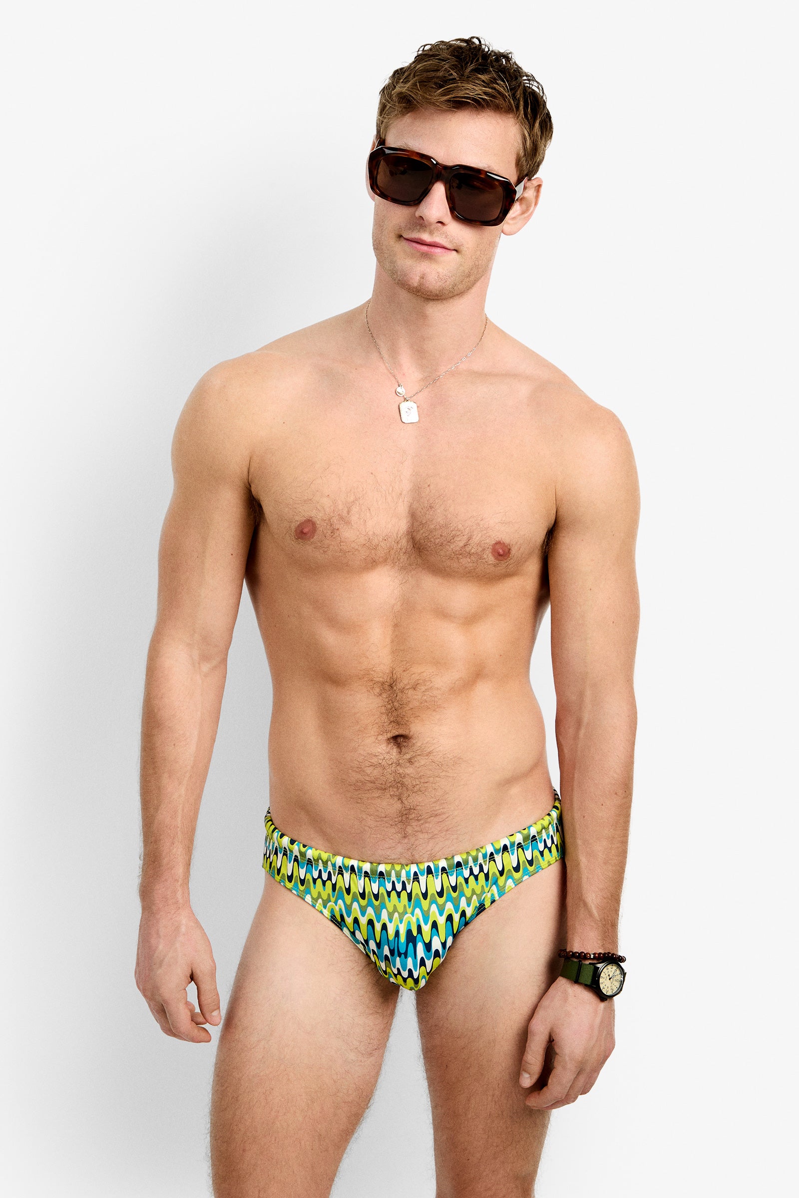 A shirtless man wearing sunglasses, a necklace, and a watch models the Lagos Swim Brief - Psychedelic Voyage against a plain white background.