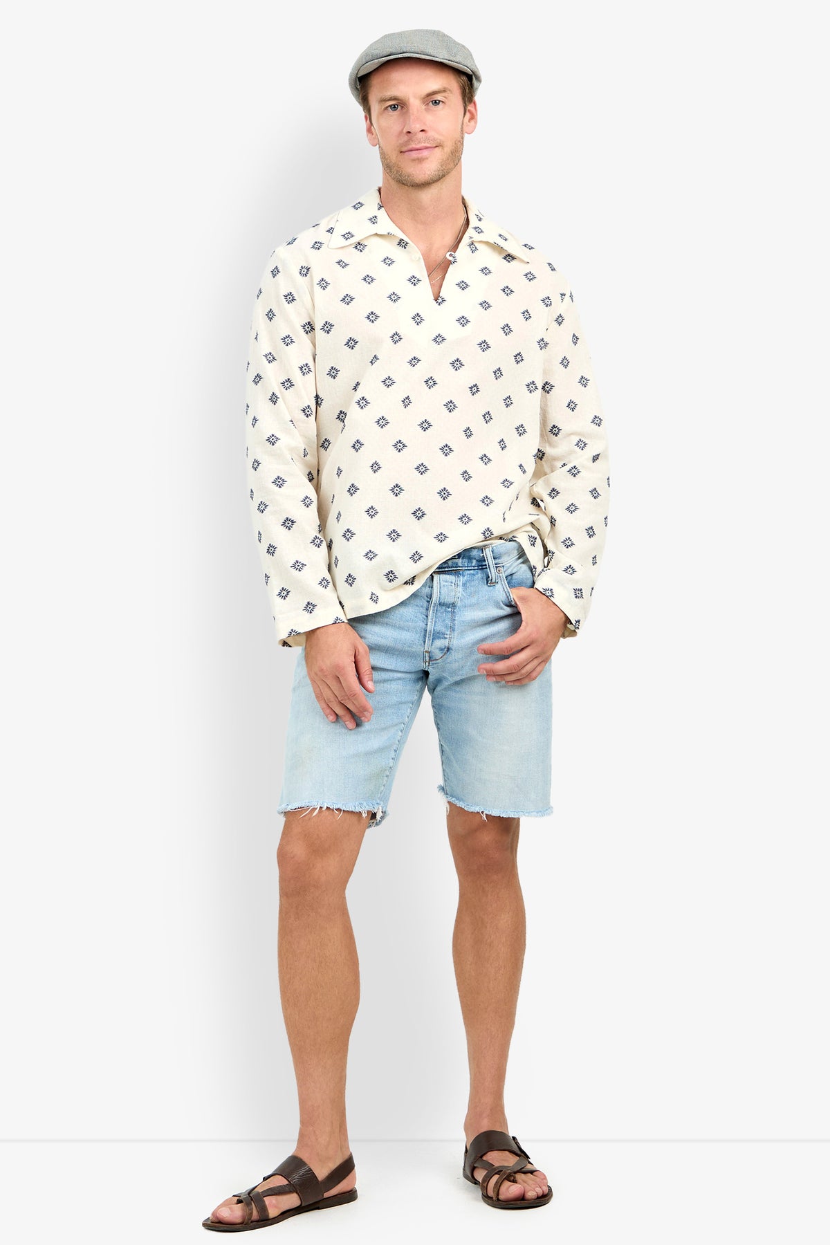 Man in a flat cap, Hendrix Long-Sleeve Pullover, light denim shorts, and brown sandals stands facing forward with a relaxed expression against a plain background.