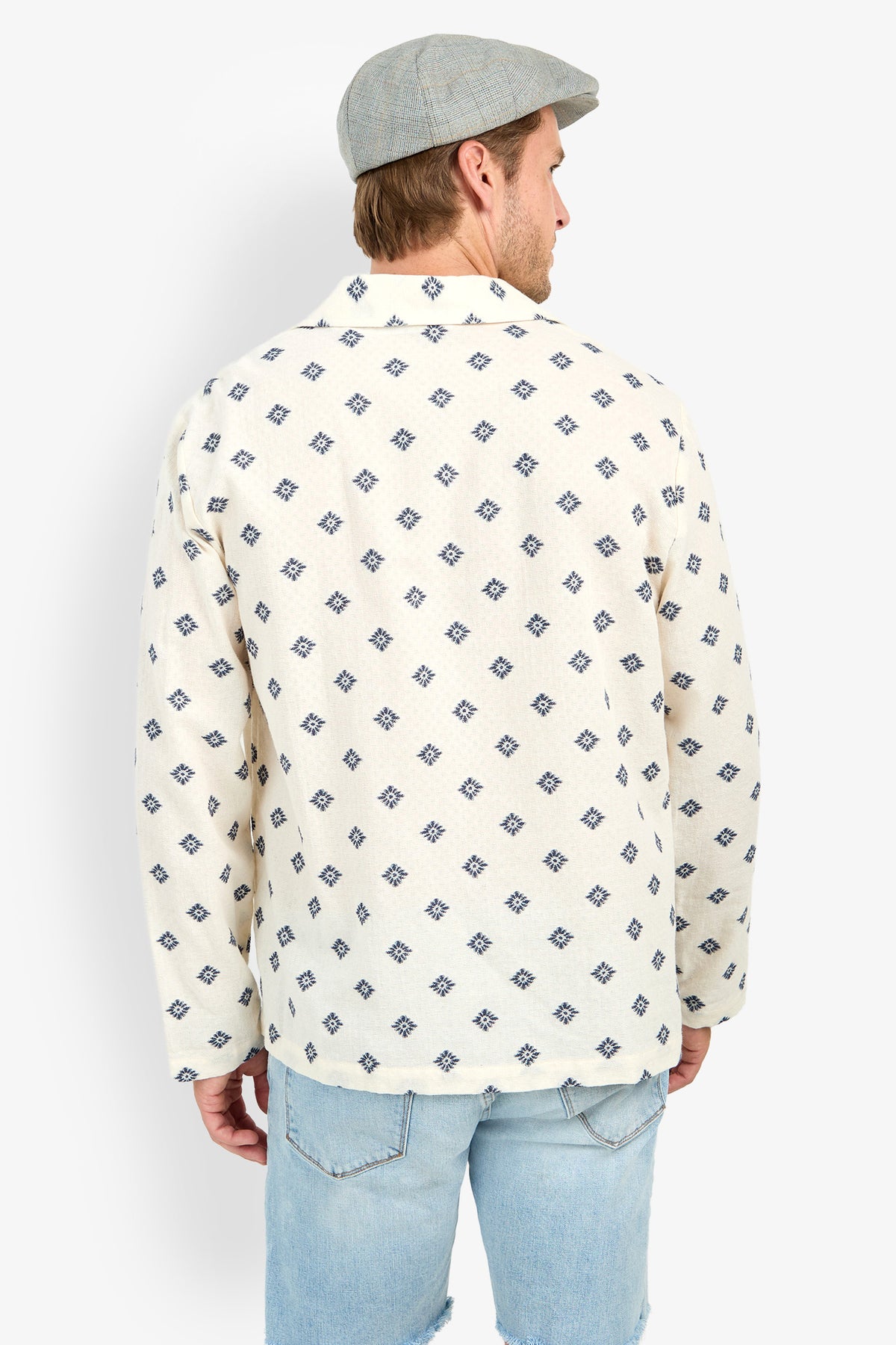 A person stands with their back to the camera against a white background, wearing a gray flat cap, Hendrix Long-Sleeve Pullover, and light blue ripped jeans.