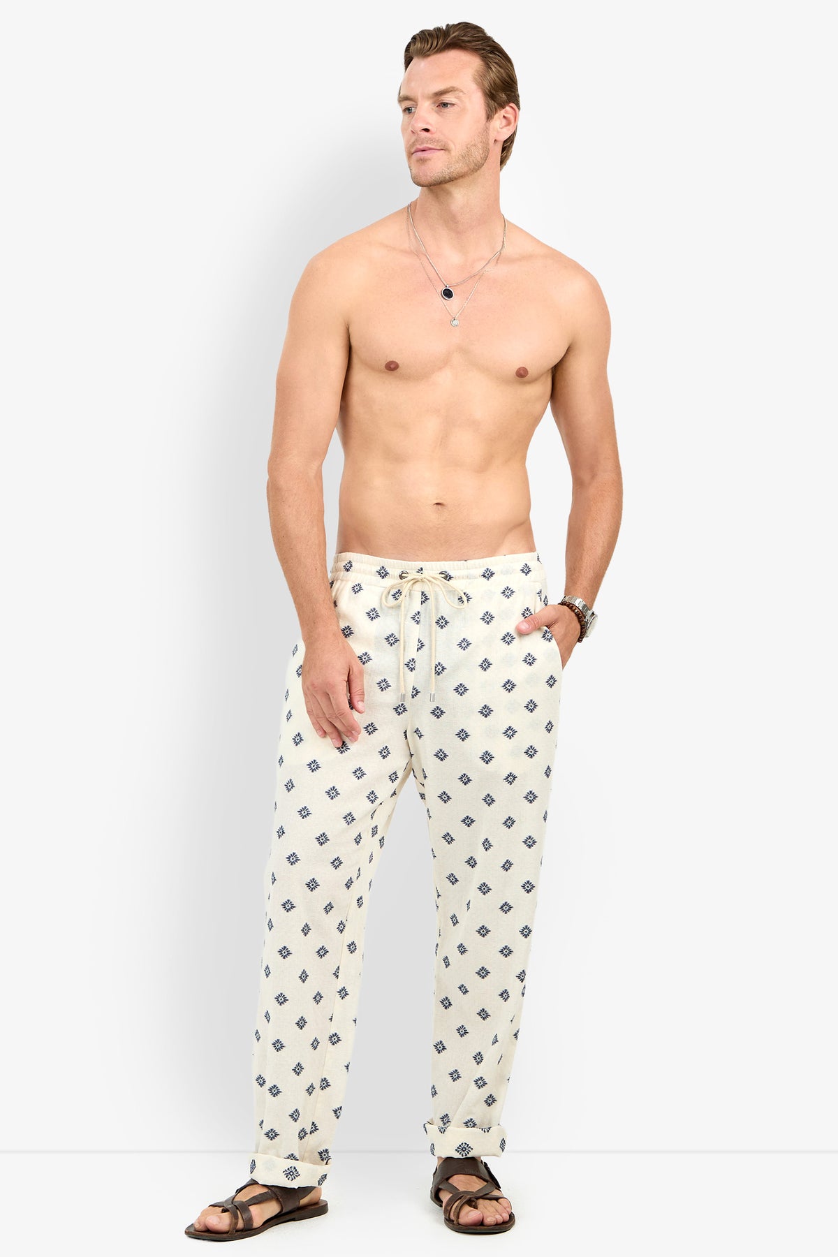 A shirtless man wears Felipe Drawstring Pant in white with a blue pattern, paired with brown sandals and necklaces, standing against a plain white background and looking to his left.