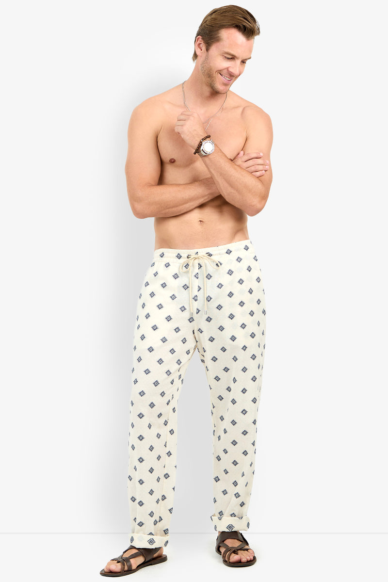 A shirtless man wearing Felipe Drawstring Pant in white and brown sandals stands smiling with his arms crossed, looking to the side.