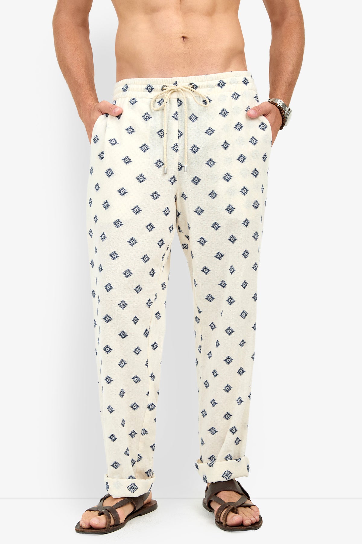 A man wearing the Felipe Drawstring Pant in cream with a blue geometric print, hands in pockets, shirtless, and paired with brown sandals.