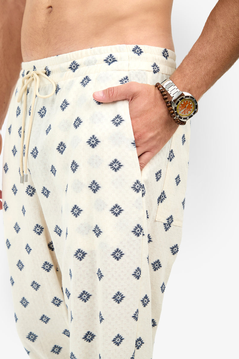 A person wears the Felipe Drawstring Pant in cream with a blue geometric pattern, hand in pocket, accessorized with a watch and beaded bracelet. Only the lower torso and arm are visible.