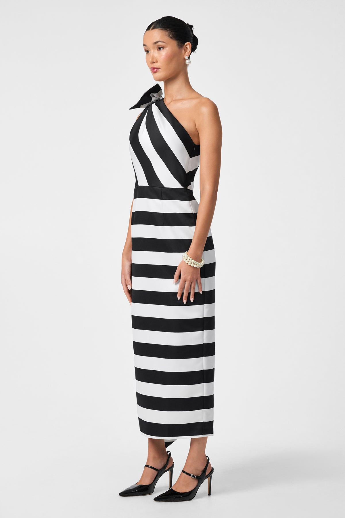 A woman stands against a plain white background wearing the Romance Dress—a black-and-white, one-shoulder, ankle-length dress with bold stripes—paired with black heels, pearl bracelets, and a sleek bun.