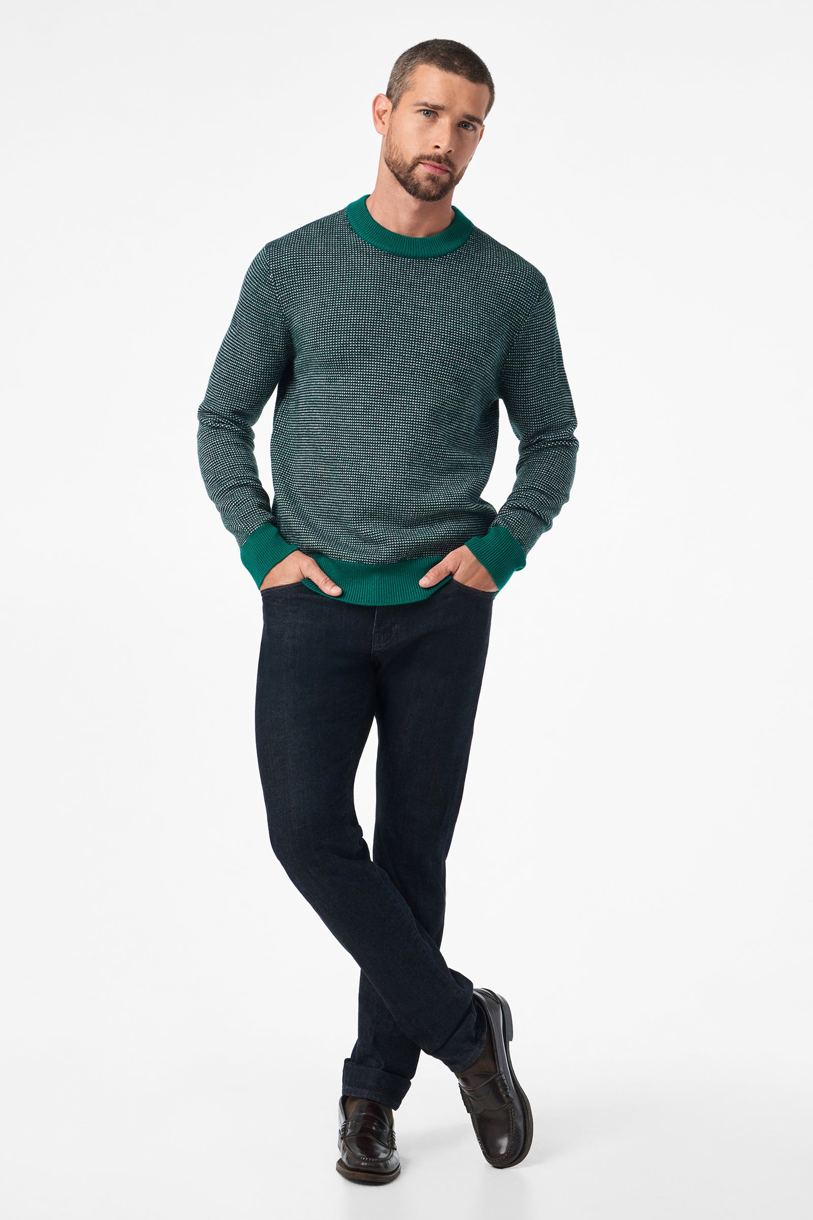 A man with short hair and a trimmed beard poses with his hands in his pockets, wearing the Ezra Crewneck in green, dark jeans, and dark shoes against a plain white background.