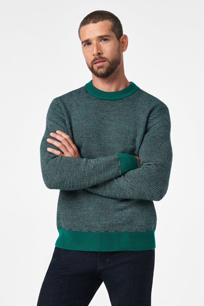 A man with short hair and a beard stands with his arms crossed, wearing the Ezra Crewneck—a green and black patterned sweater with matching green cuffs, collar, and hem—paired with dark jeans against a plain white background.