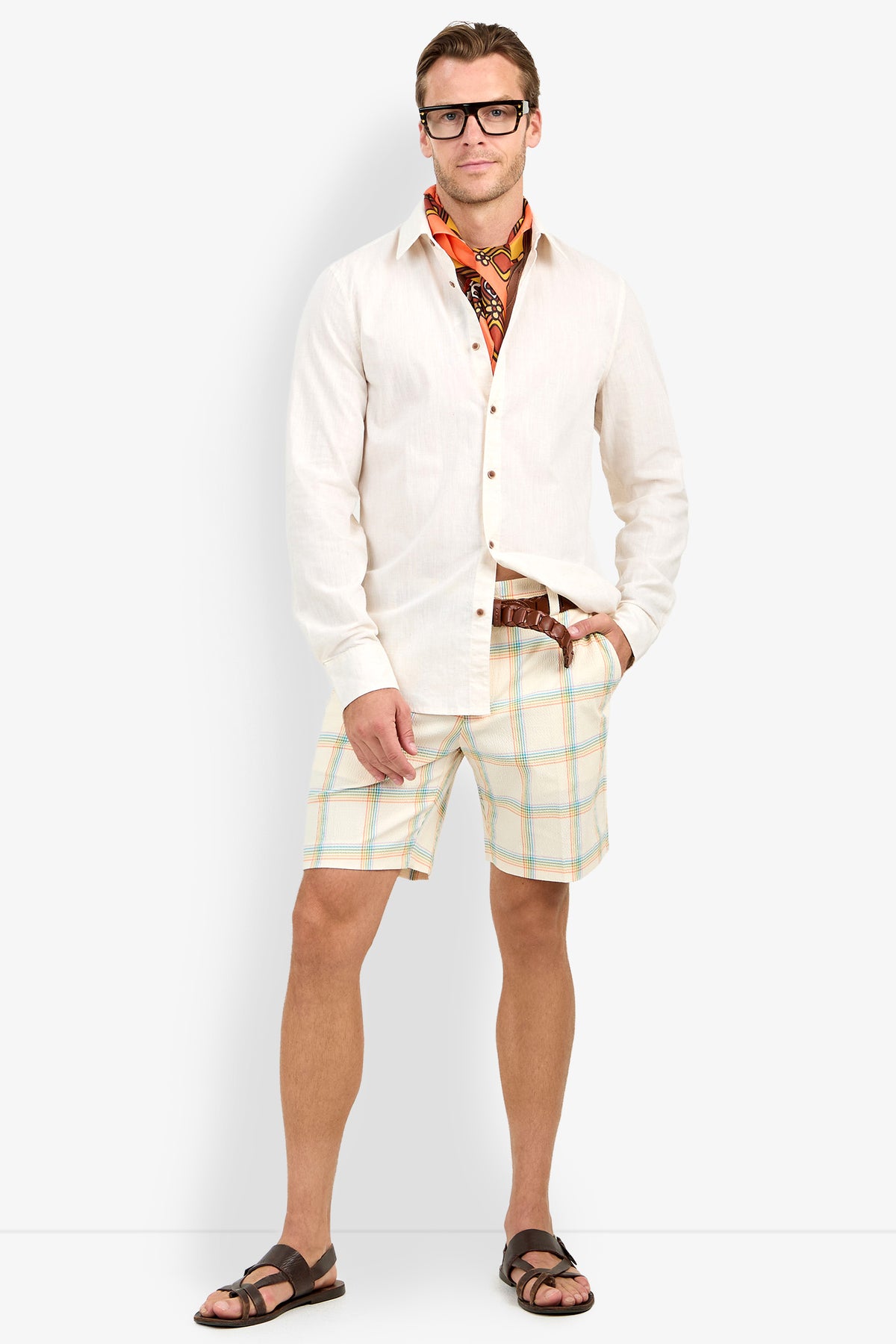 Man wearing glasses, the Aaron Shirt (white button-up), orange patterned scarf, plaid shorts, brown belt, and brown sandals stands casually with one hand in pocket against a plain white background.