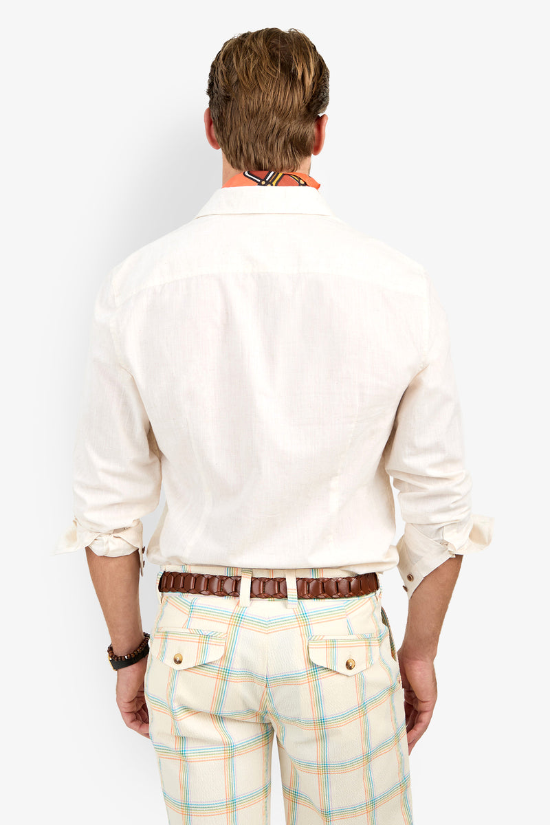 A man stands facing away, wearing the Aaron Shirt with rolled-up sleeves, plaid pastel-striped pants, a brown belt, bracelet, and a colorful scarf around his neck.