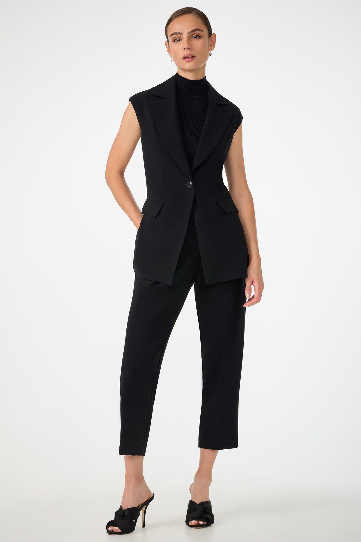 A woman stands against a plain background wearing the Boabab Sleeveless Jacket over a black turtleneck, paired with cropped black pants and black heeled mules. Her hair is pulled back, and she poses with one hand in her pocket.