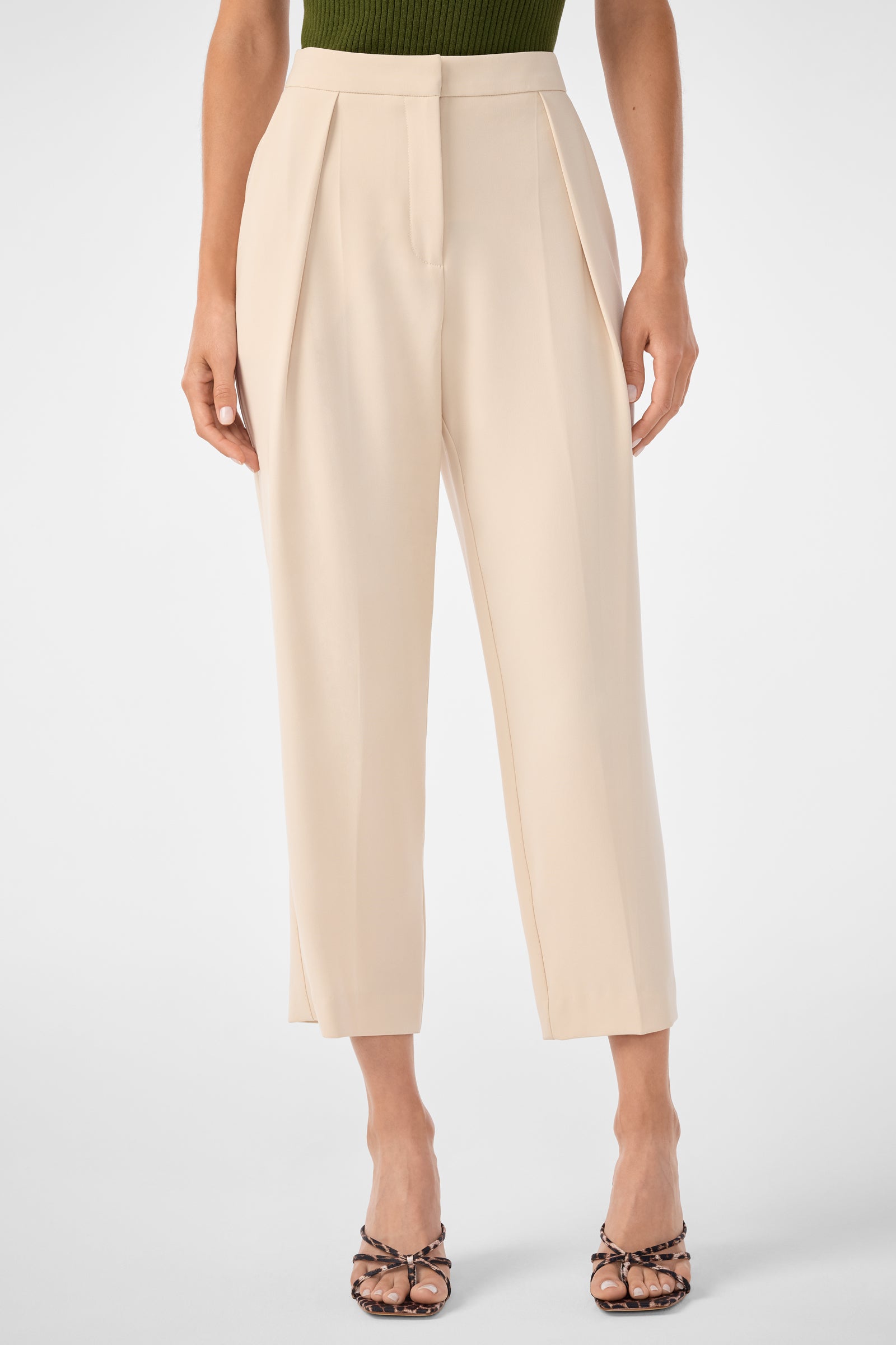 Someone in the Larch Tailored Cropped Pants in Poplar, high-waisted and pleated with a front zipper and button, styled with a green top and open-toe heeled sandals.