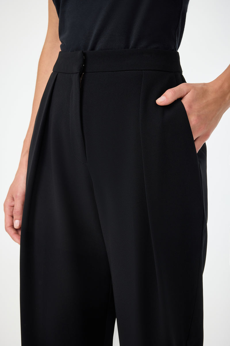 A person wears Larch Tailored Cropped Pants and a black top, standing against a plain background with one hand in their pocket and the other arm relaxed by their side.