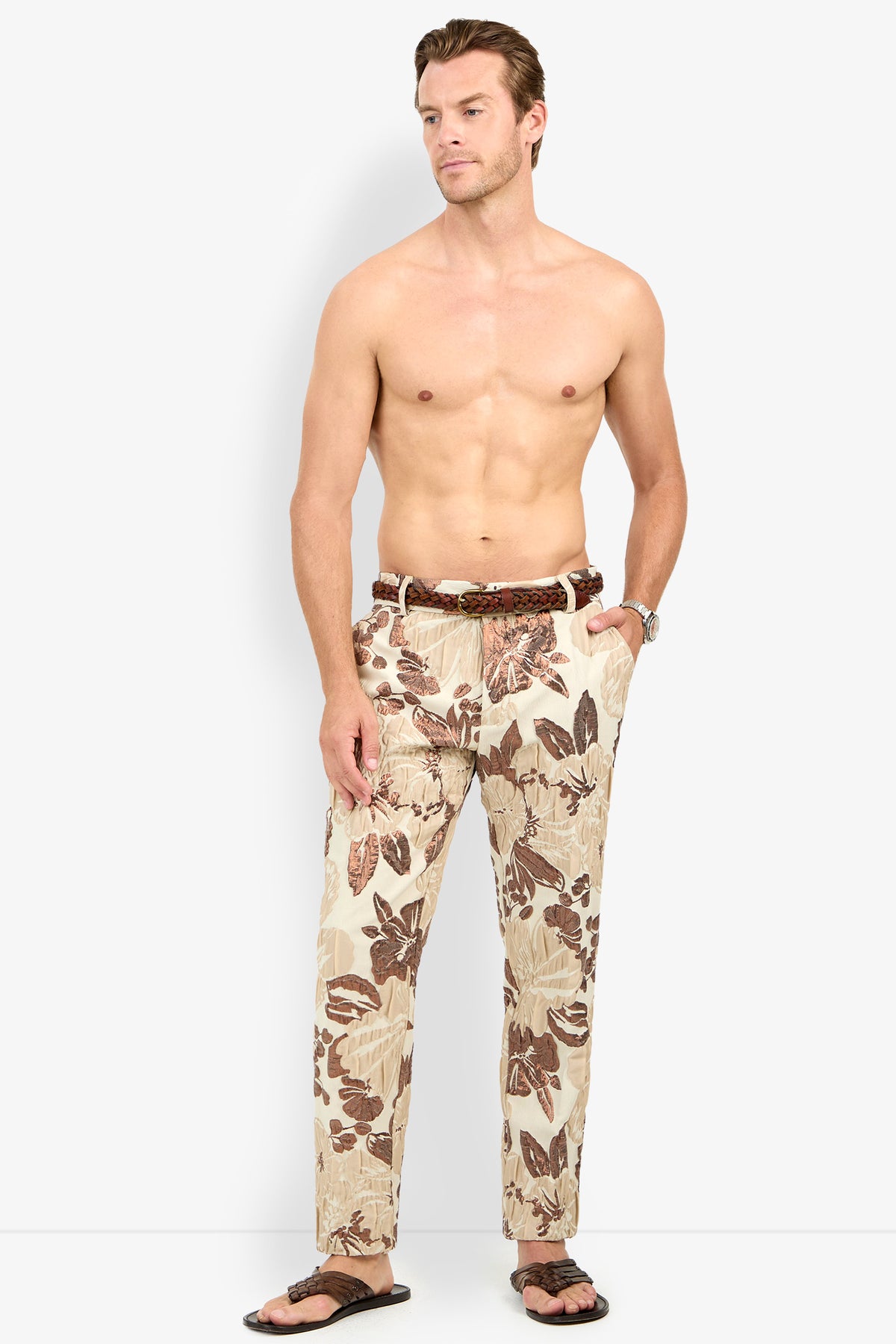 A shirtless man wears Clyde Slim Trouser in beige and brown floral print, paired with a belt, flip-flops, and a watch, standing against a plain white background with hands in his pockets.