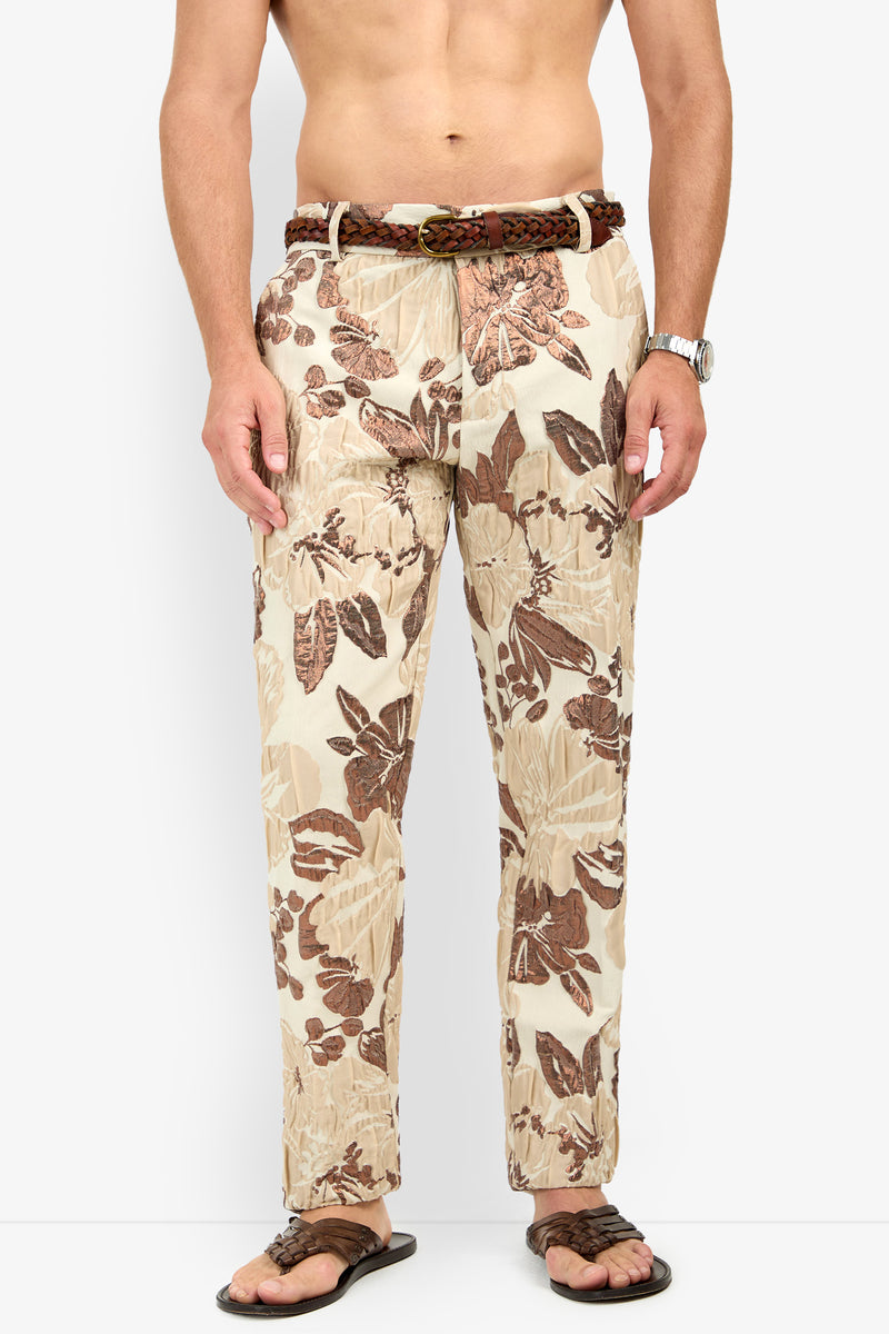 A shirtless man wearing the Clyde Slim Trouser—beige pants with a brown tropical leaf pattern—paired with a woven brown belt, watch, and brown sandals, stands against a plain white background.