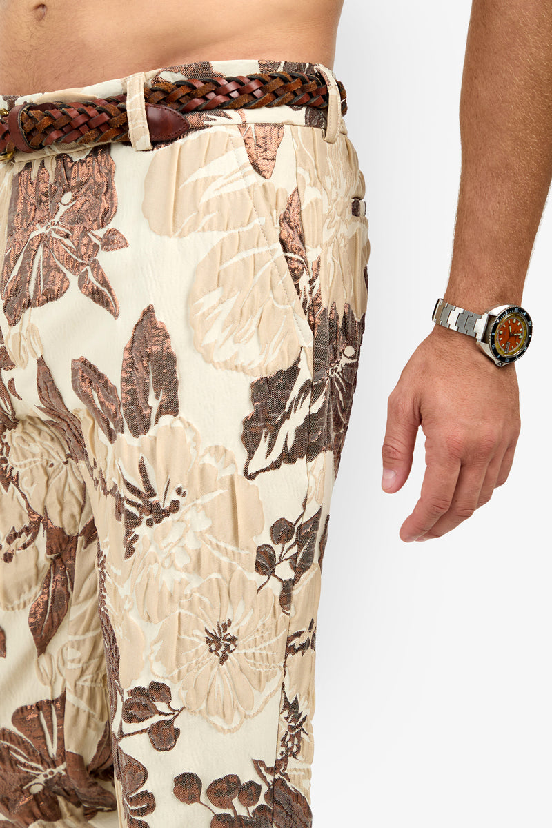 A person wears Clyde Slim Trouser in beige and brown floral print, paired with a brown woven belt and a silver wristwatch with an orange face. Only the torso and right arm are shown on a white background.