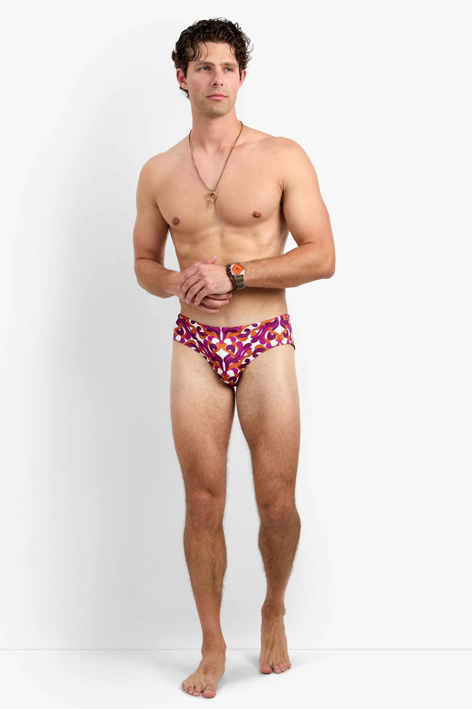 A man with short curly hair stands barefoot against a white background, wearing the Bondi Swim Brief - Dewdrop, along with a necklace and wristwatch. He looks slightly to the side with a neutral expression.