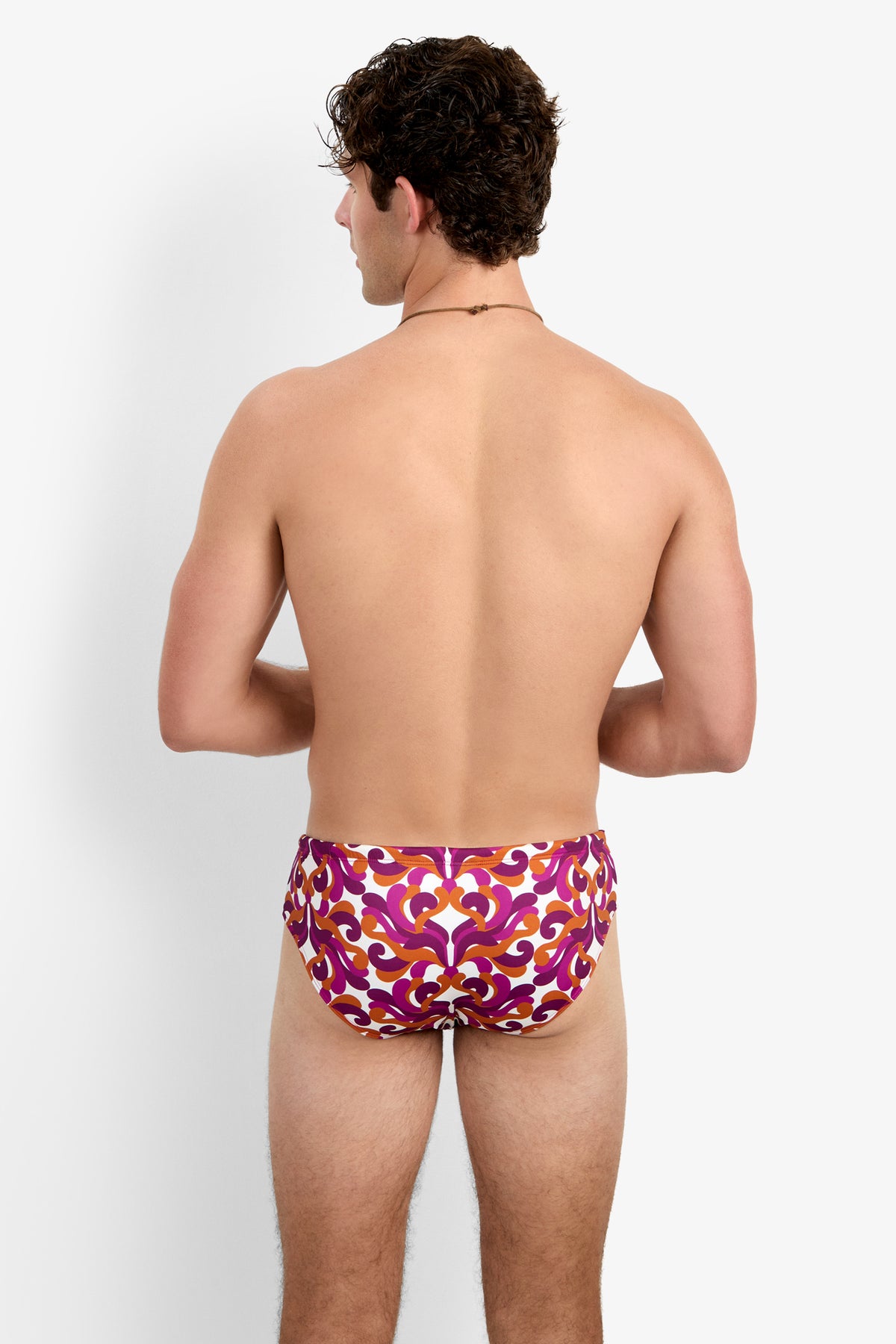 A shirtless man faces away, wearing the Bondi Swim Brief - Dewdrop featuring a red, orange, and purple ornate pattern. The background is plain white.