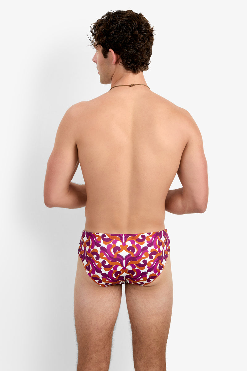 A shirtless man faces away, wearing the Bondi Swim Brief - Dewdrop featuring a red, orange, and purple ornate pattern. The background is plain white.