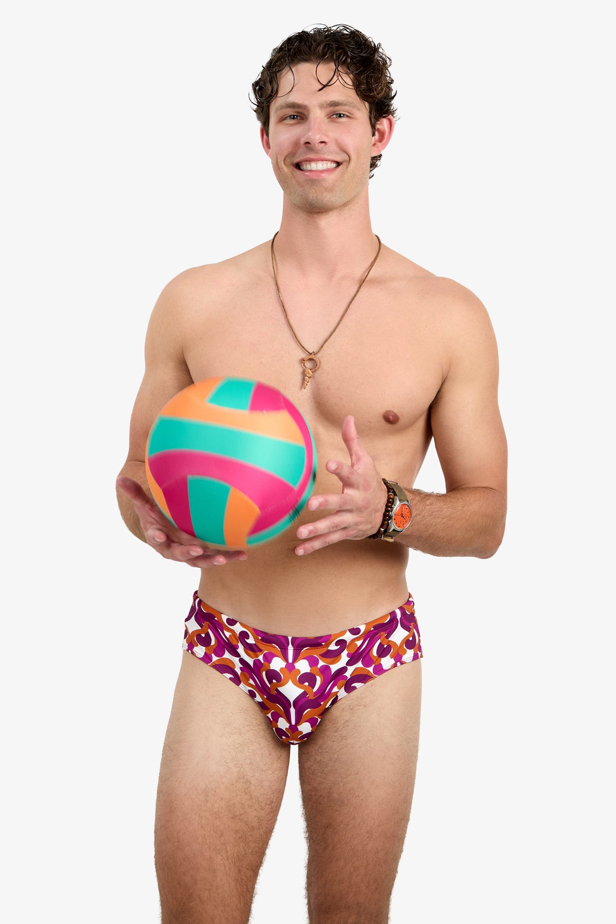 A smiling man wears the Bondi Swim Brief - Dewdrop as he holds a colorful volleyball. He accessorizes with a necklace and watch, standing against a plain white background.