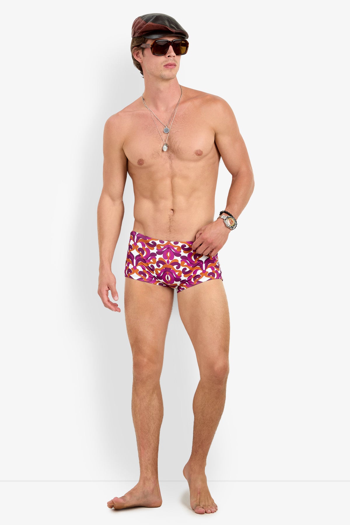 A shirtless man models the Jet Set Swim Trunk, paired with sunglasses, a cap, a wristwatch, and necklaces, standing barefoot against a plain white background.