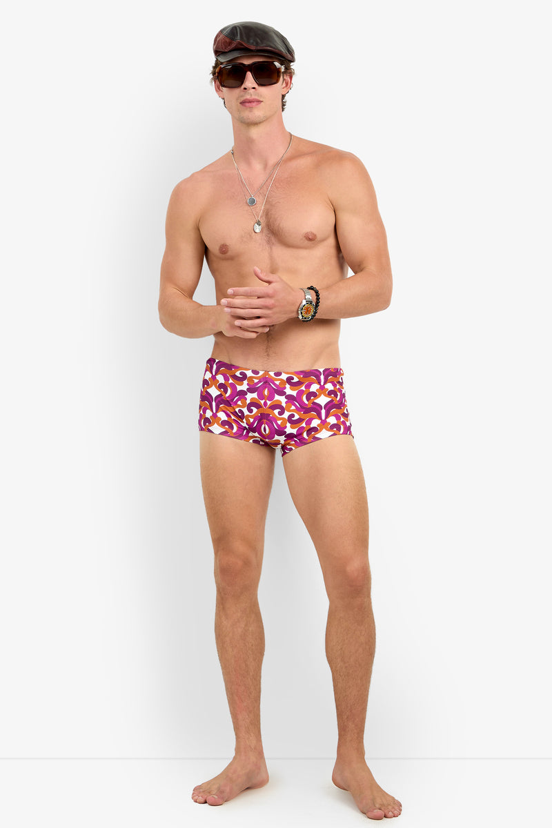 A shirtless man wears Jet Set Swim Trunk, sunglasses, a black cap, and layered necklaces and bracelets while standing barefoot against a white background.