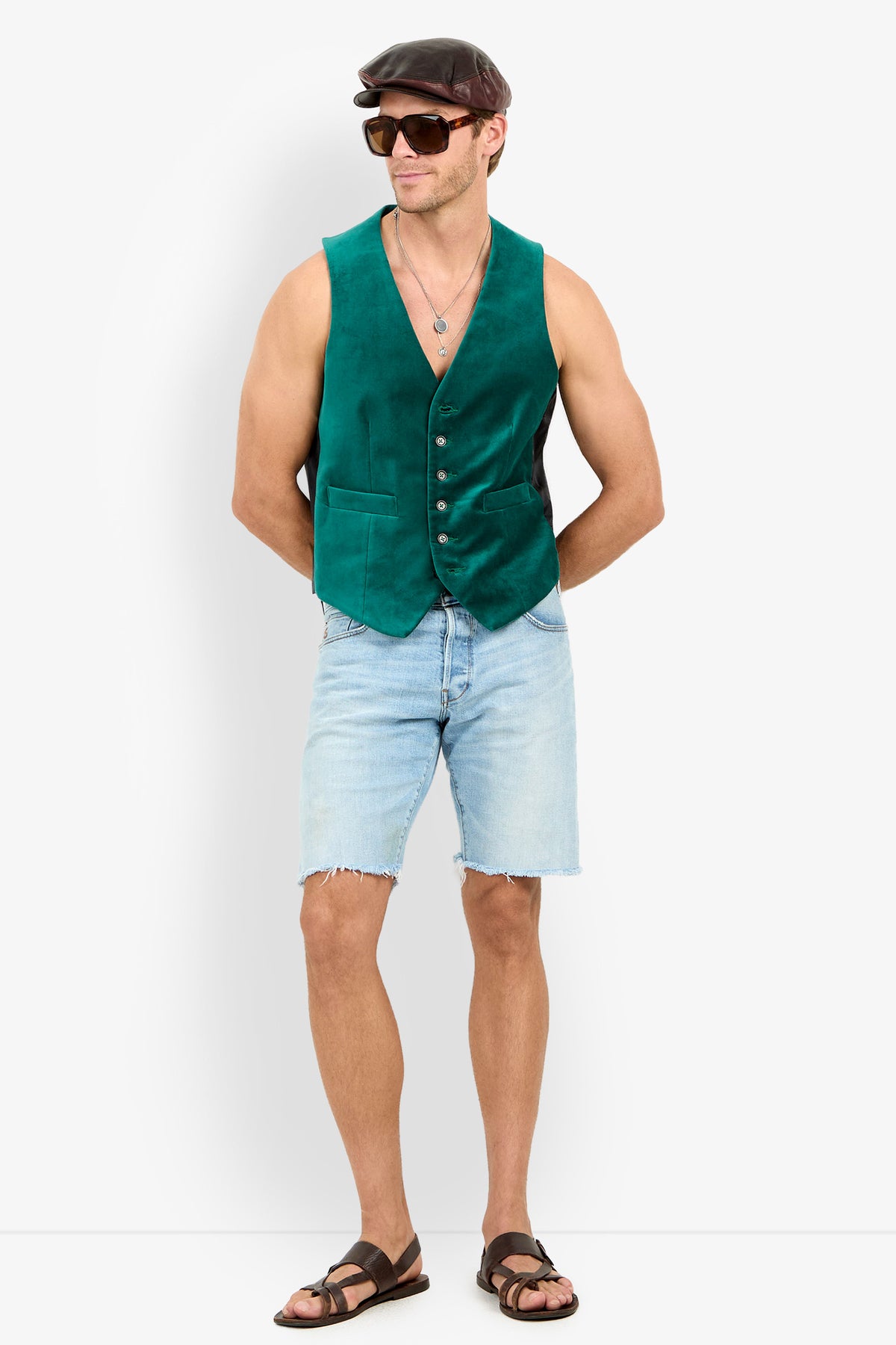 A man in the Irving Velvet Vest, light denim shorts, brown sandals, a brown flat cap, sunglasses, and a necklace stands with his hands behind his back against a white background.