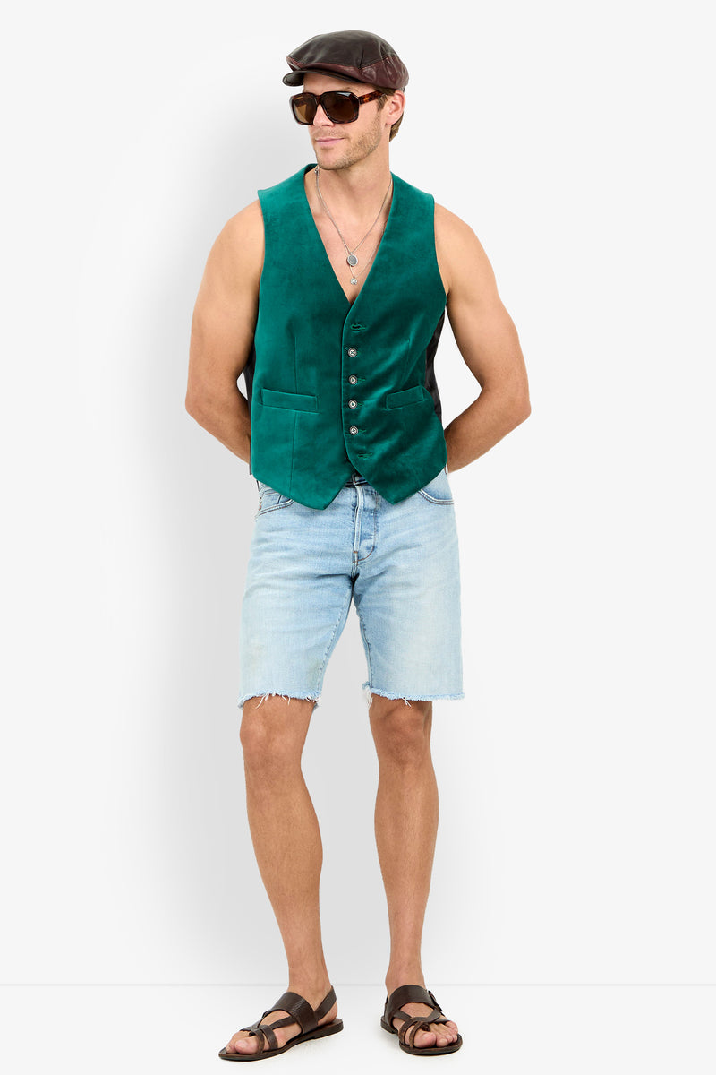 A man in the Irving Velvet Vest, light denim shorts, brown sandals, a brown flat cap, sunglasses, and a necklace stands with his hands behind his back against a white background.