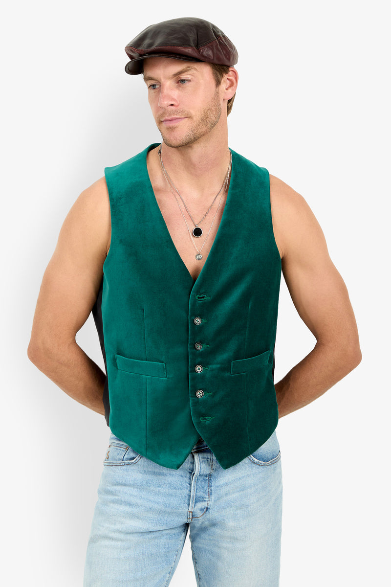 A man in an Irving Velvet Vest, blue jeans, layered necklaces, and a brown flat cap stands with hands behind his back, looking to the side.