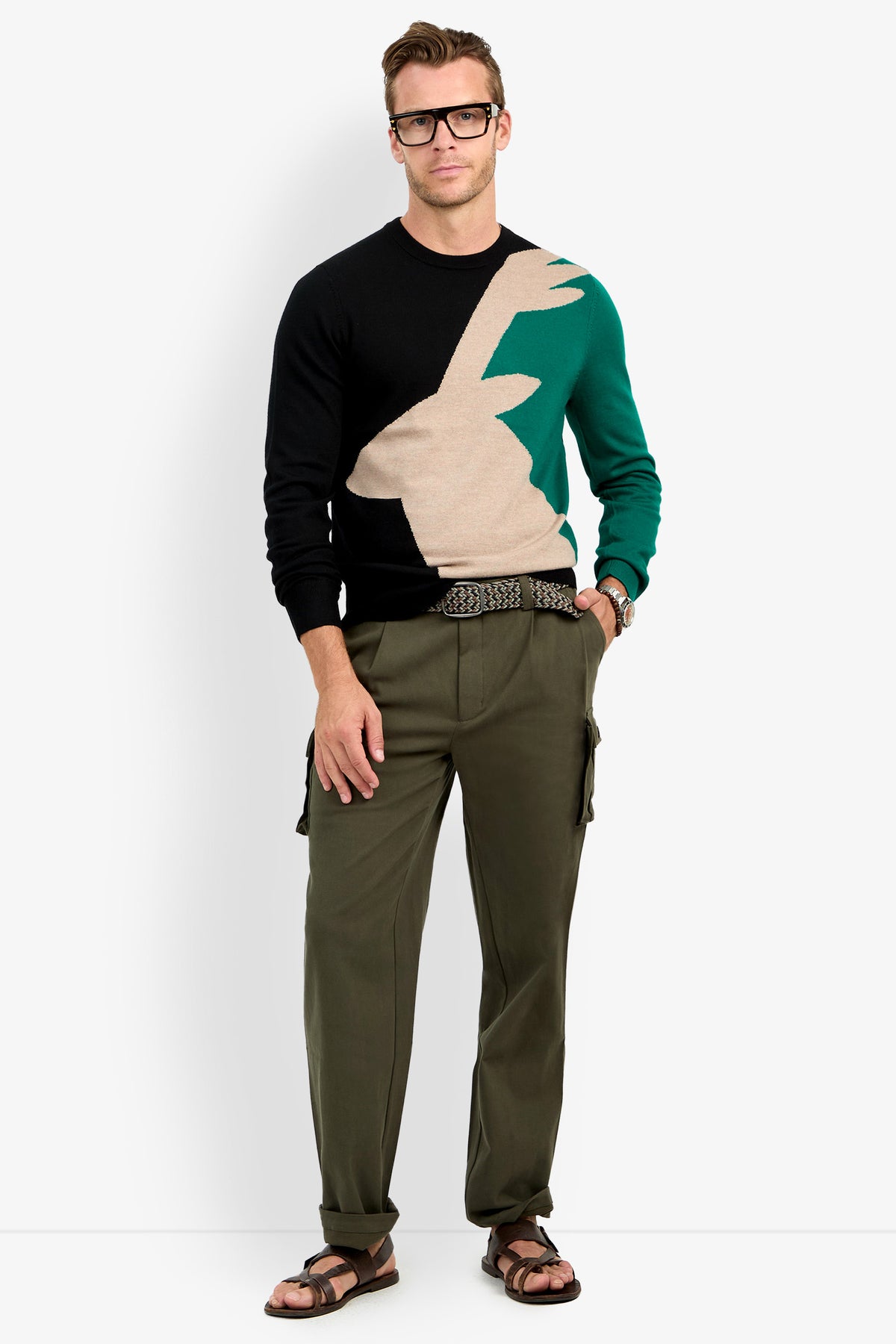 A man stands facing forward against a white background, wearing glasses, a Crewneck Sweater with a large graphic, olive cargo pants, a patterned belt, and brown sandals.