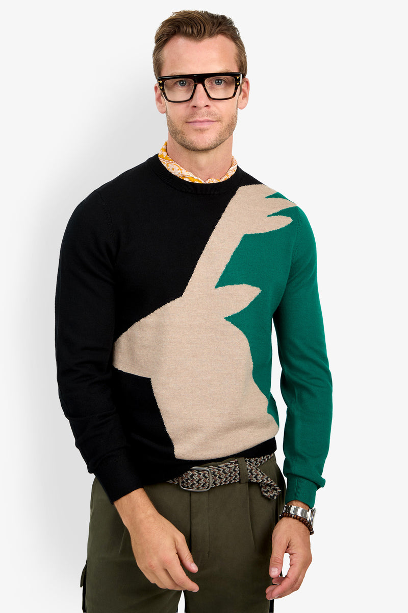 A man in thick black glasses, olive green pants, a patterned belt, and the Crewneck Sweater—featuring a multicolored abstract beige and green design—stands against a plain white background.