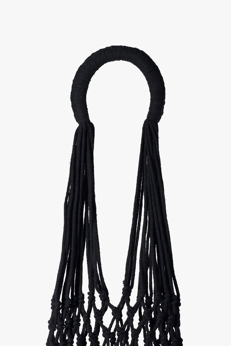 The Nannacay Kim Woven Bag features a black circular handle wrapped in fabric, with long, loose black cords hanging down to form the upper section of the intricately knotted bag, all set against a white background.