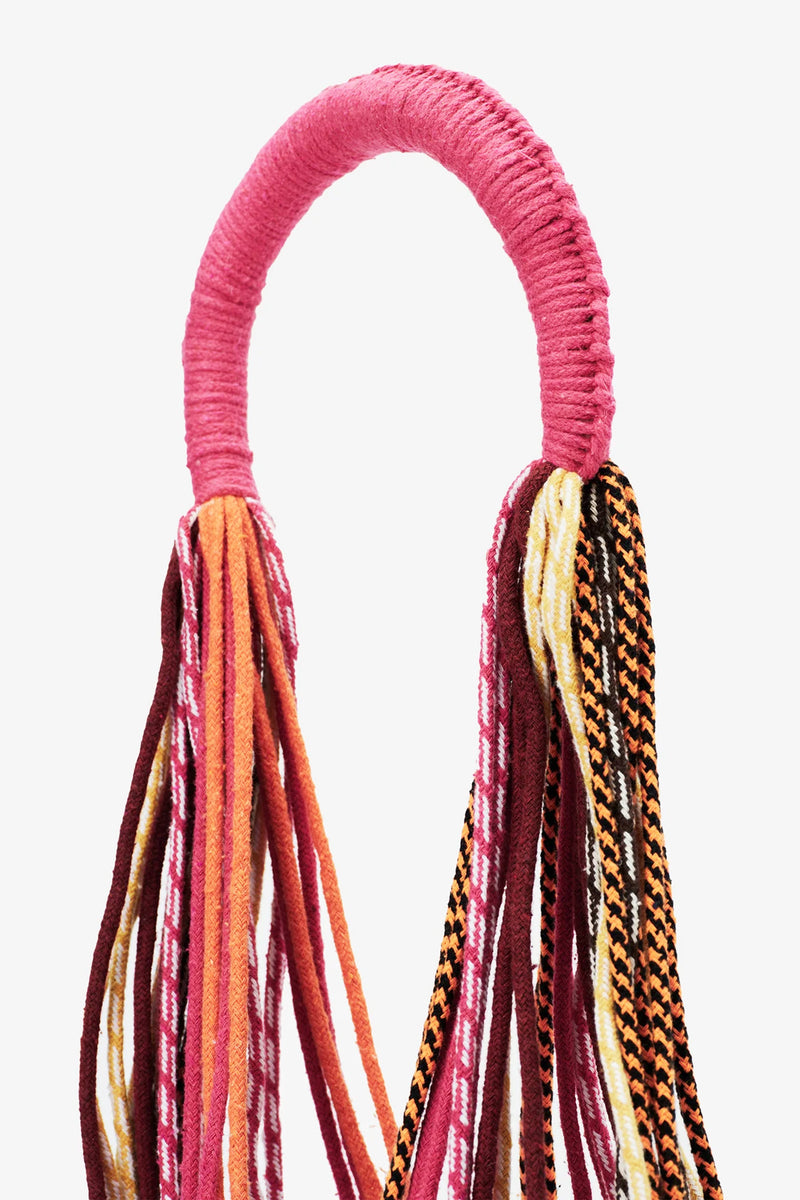 The Nannacay Kim Woven Bag features a pink fabric-wrapped handle with multiple long, colorful woven textile strands hanging down, showcasing patterns in red, pink, orange, white, and black.