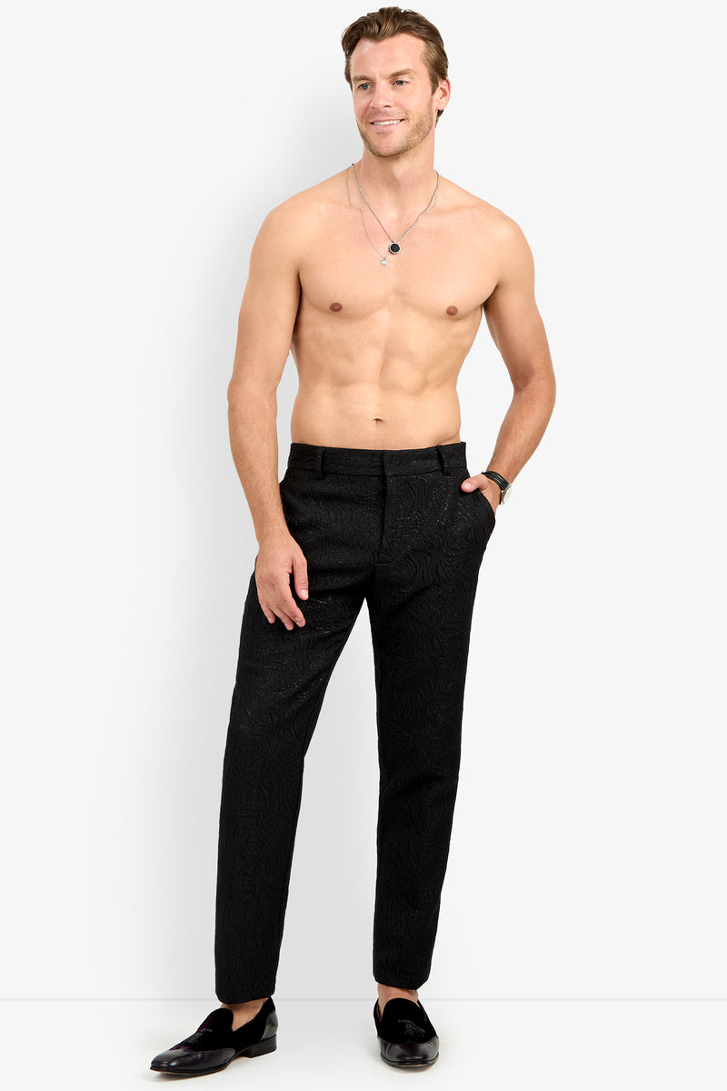 A shirtless man stands against a white background, wearing the Clyde Slim Trouser in black, paired with black loafers, a watch, and two necklaces. He is smiling with one hand in his pocket.