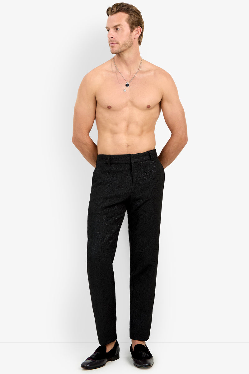 A shirtless man stands against a plain white background, hands in pockets, wearing Clyde Slim Trouser and black loafers. He has short, slicked-back hair and layered necklaces.