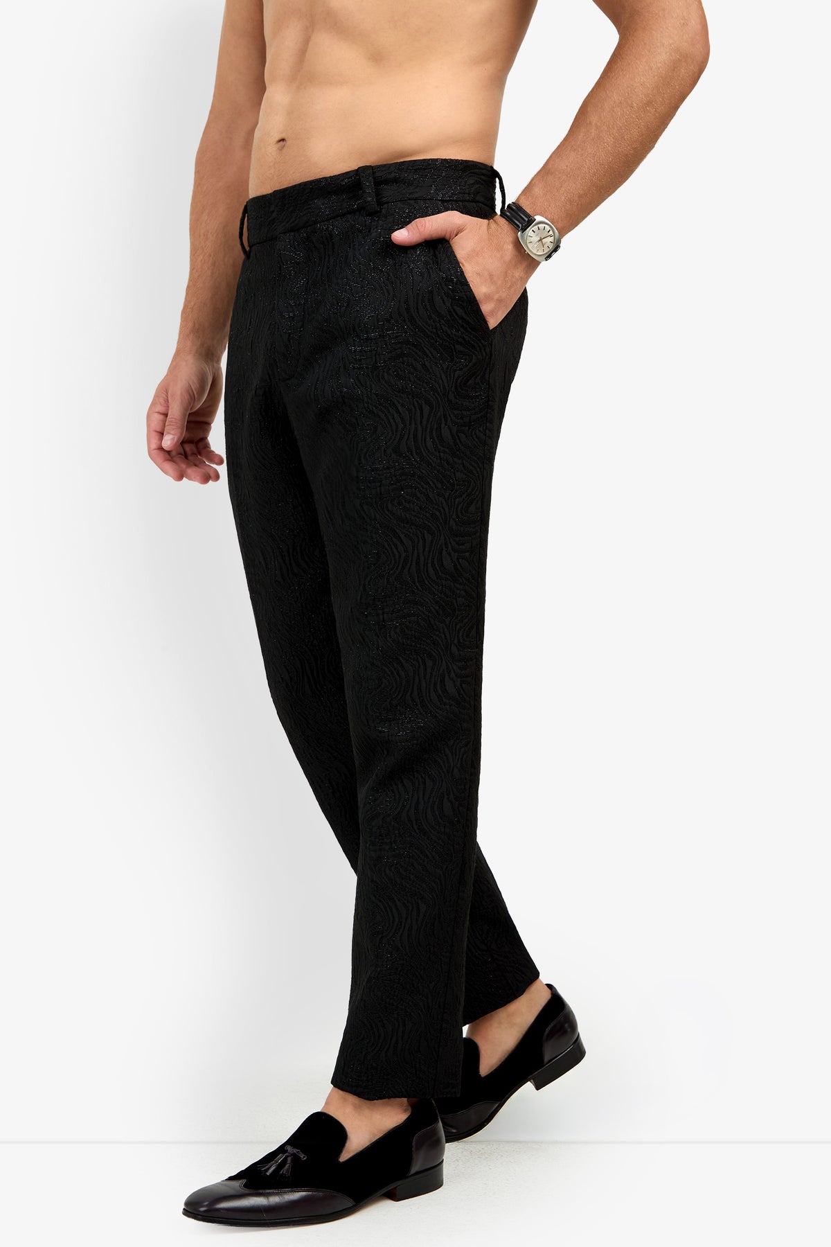 A shirtless man wearing Clyde Slim Trouser, black loafers with tassels, a wristwatch, and his hand in his pocket stands against a plain white background.