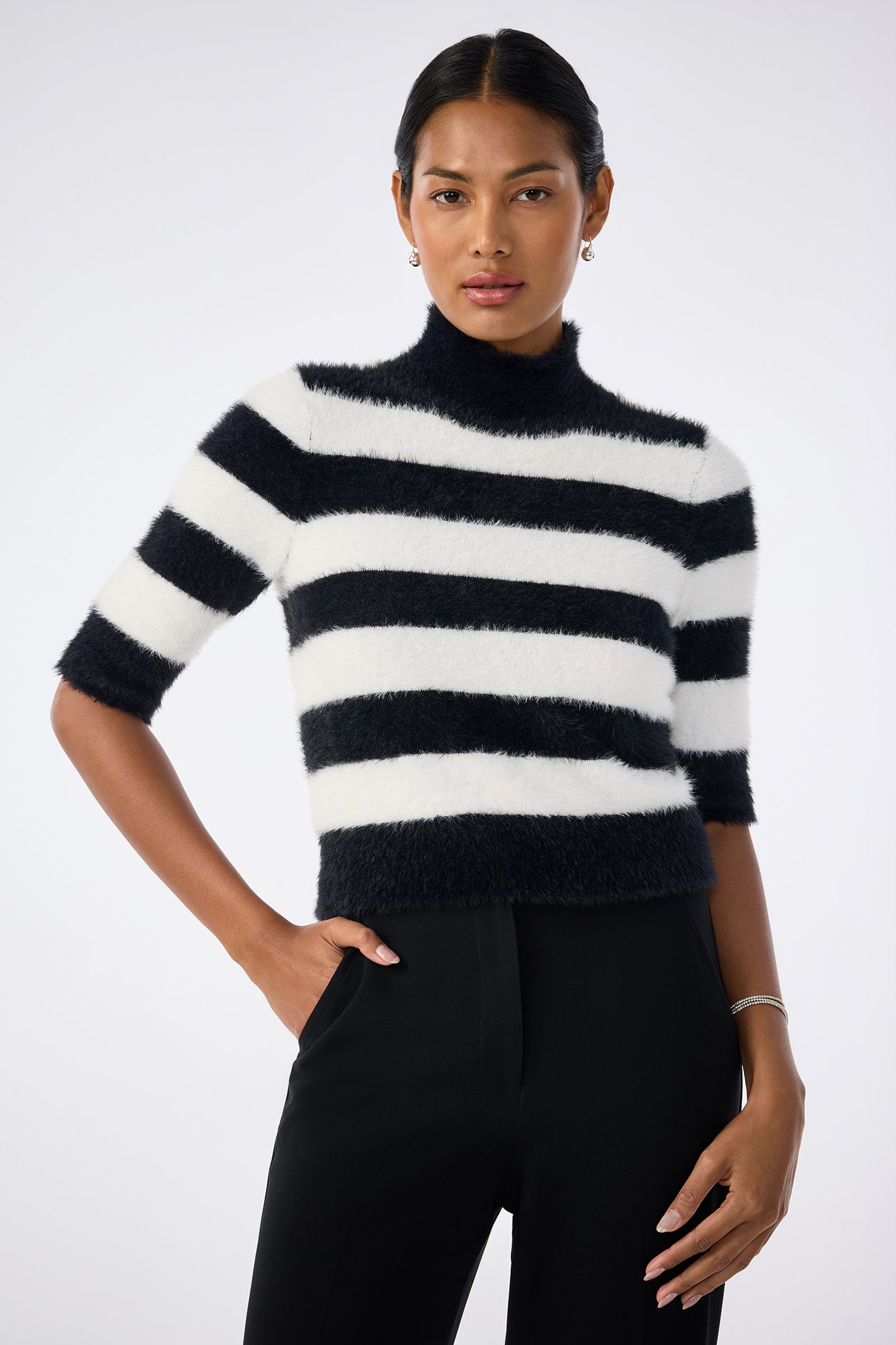 A woman poses confidently against a plain light background, wearing the Mali Mock Neck Sweater—a fuzzy black and white striped short-sleeve top—paired with black pants.
