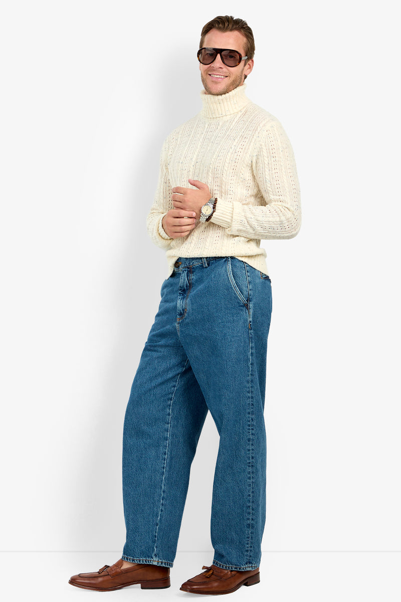 A man smiles with his arms crossed, wearing sunglasses, blue jeans, brown loafers, and a Wesley Turtleneck Sweater in front of a white background.