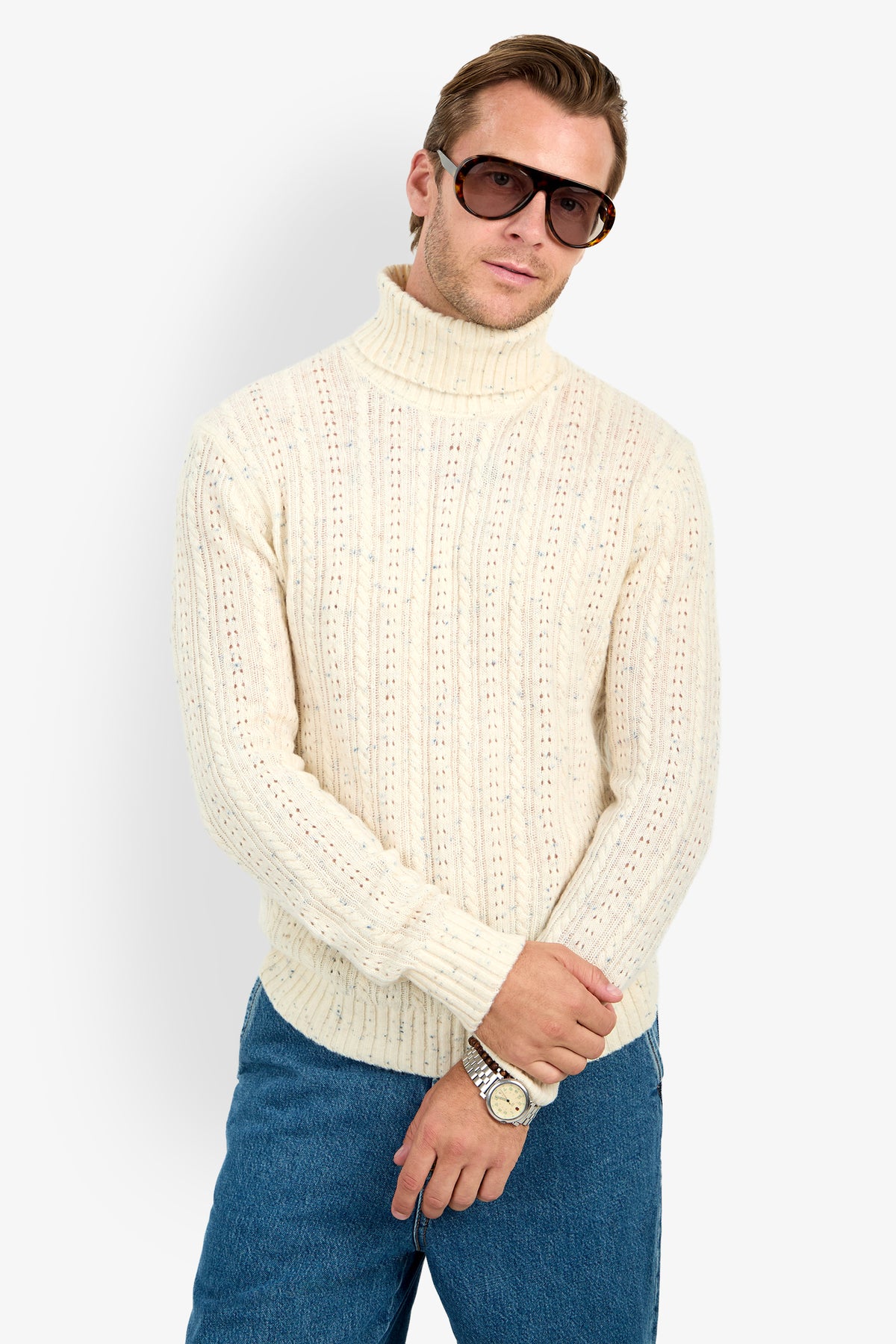 A person wearing sunglasses, the Wesley Turtleneck Sweater, blue jeans, and a wristwatch stands against a white background with hands crossed at the waist.
