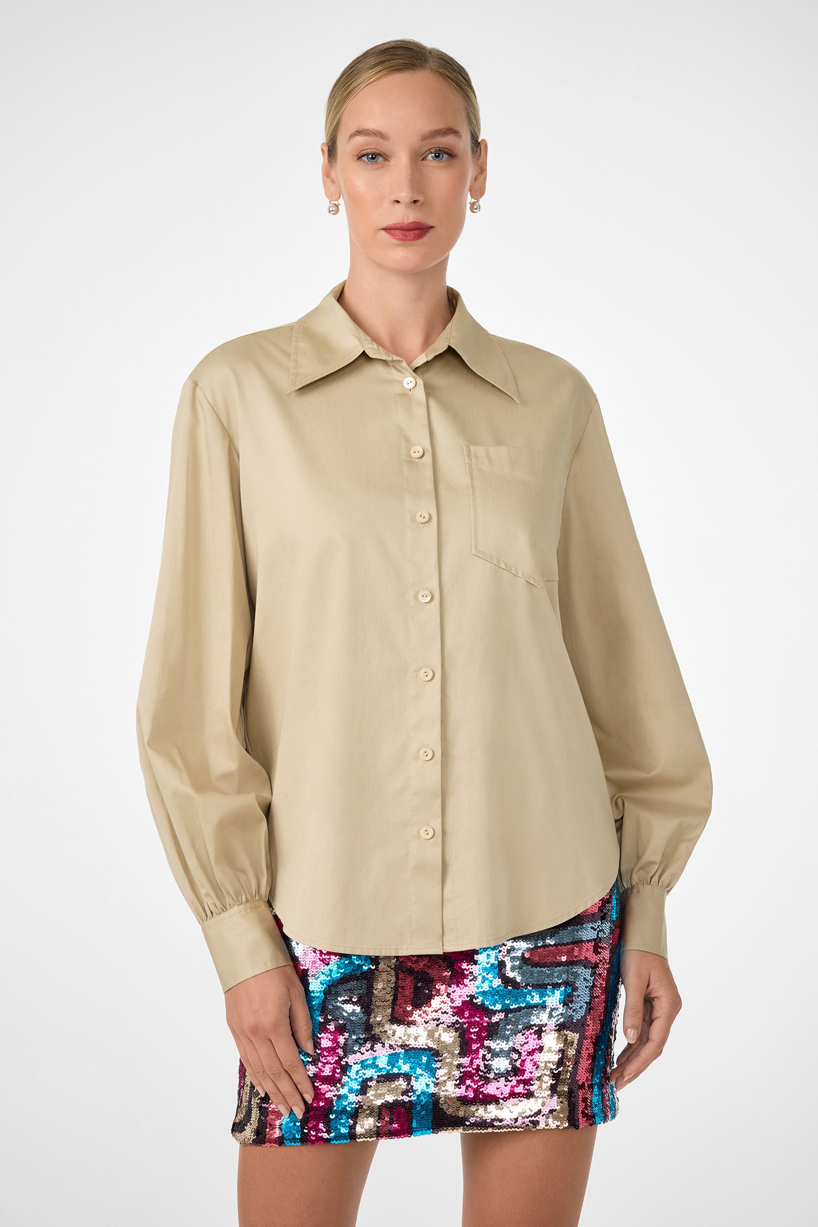 A woman wearing the Burle Top—a beige, long-sleeve, button-up shirt with a front pocket—is paired with a colorful geometric sequin mini skirt and stands facing forward against a plain white background.