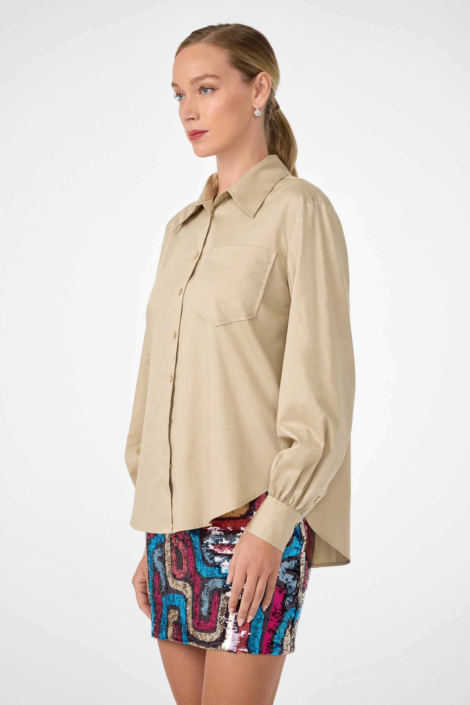 A woman in profile wears the Burle Top, a beige long-sleeve button-up shirt, paired with a colorful abstract sequin mini skirt. Her hair is tied back with subtle makeup against a plain white background.