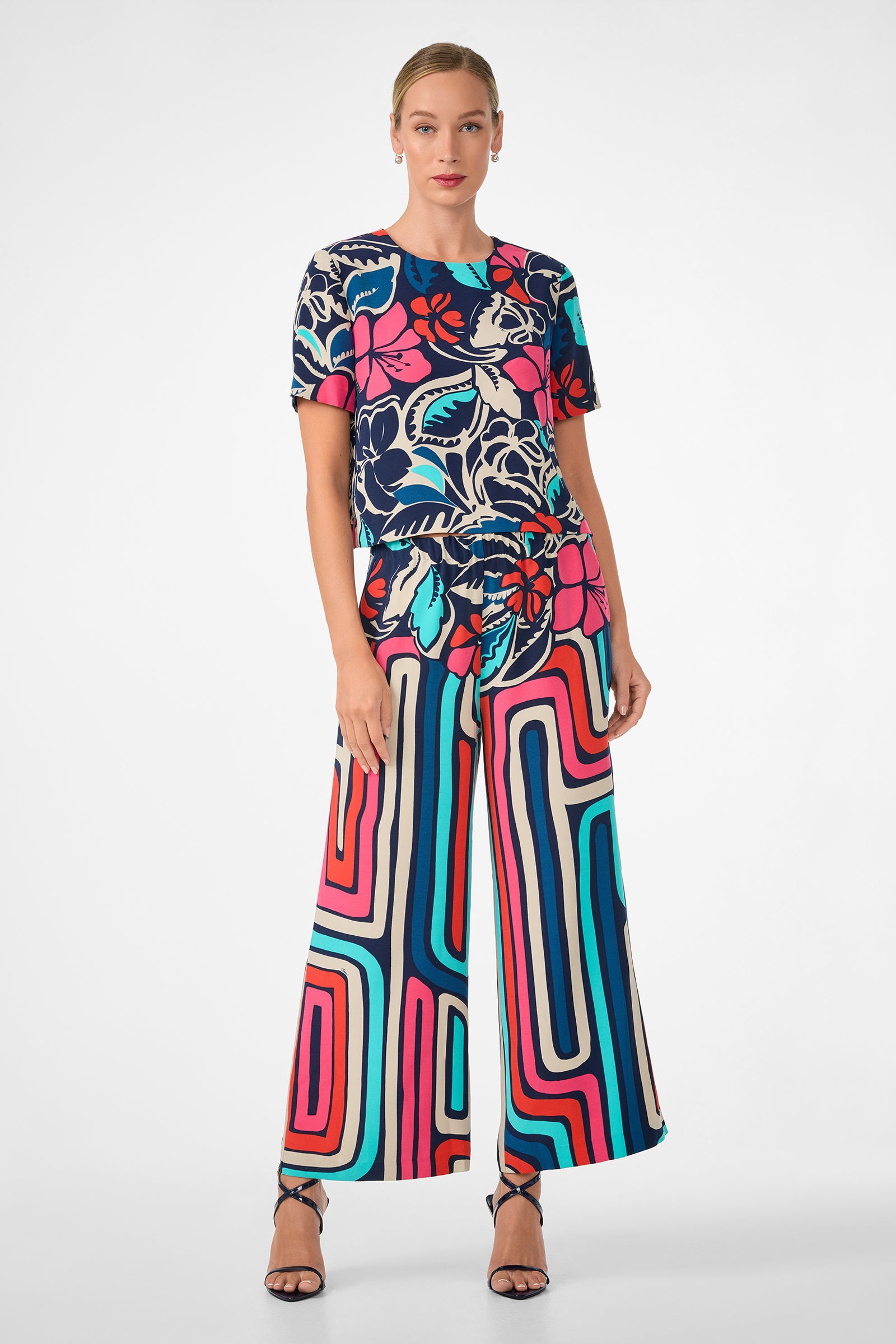 A woman wears the Sitio Top, a short-sleeve floral top, paired with wide-leg pants in bold pink, blue, red, and white geometric patterns. She completes the look with black strappy heels and has her hair pulled back.