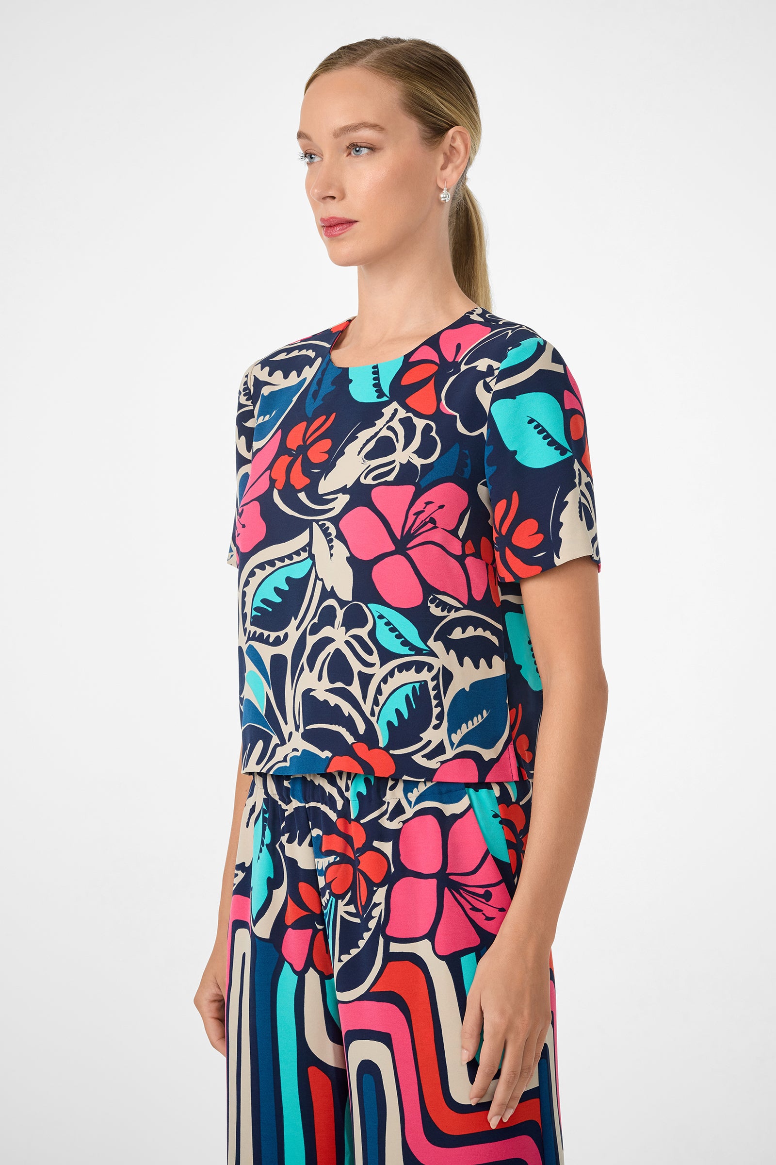 A woman with blond hair in a ponytail models the Sitio Top, a colorful piece with bold floral and geometric patterns in blue, pink, red, teal, and cream. She is shown in profile against a plain white background.