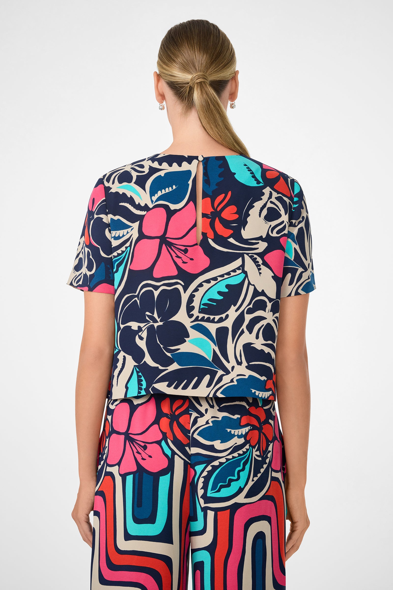 A woman faces away, wearing the Sitio Top and matching pants featuring a bold floral and geometric design in blue, red, pink, black, and turquoise. Her straight hair is styled in a low ponytail.